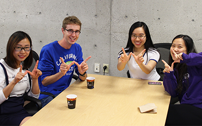Four students sitting around a table with assorted warm beverages, smiling and holding their hands in the shape of a "W"