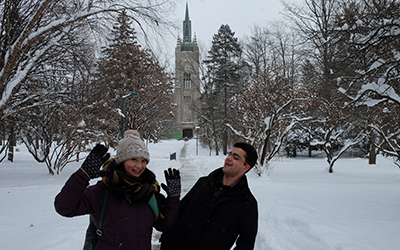 Students dressed in winter gear smiling and waving with Middlesex College in the background. Lots of snow and looks very cold.